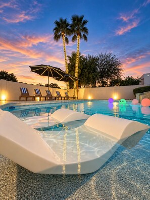 Une piscine entourée de palmiers, avec des chaises longues et un parasol, le tout baigné par la lumière d’un coucher de soleil.