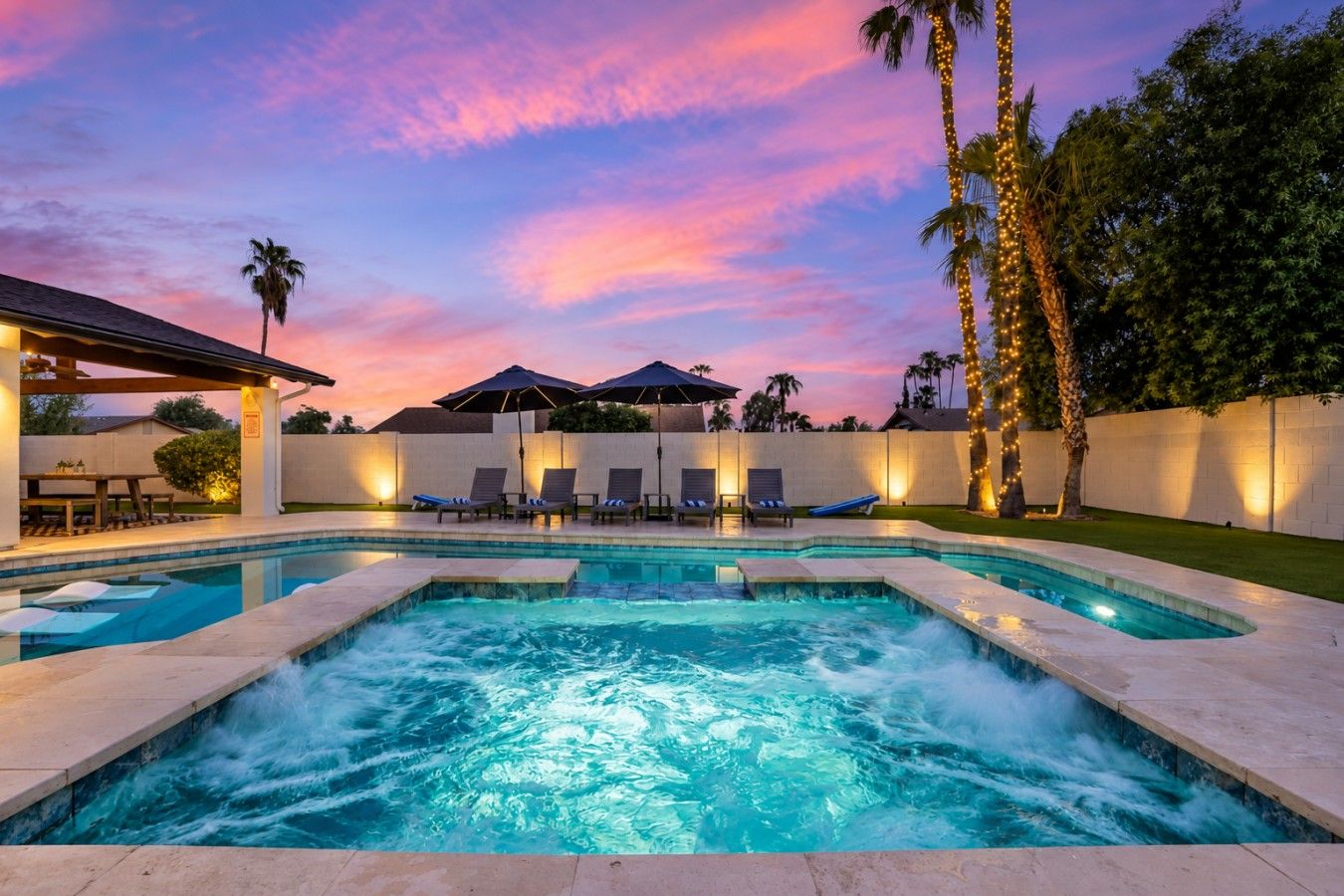 A swimming pool with lounge chairs and umbrellas, surrounded by palm trees and a sunset sky.