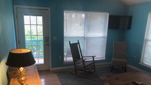 A living room with a wooden table, a lamp, a rocking chair, a sofa, and a TV mounted on the wall. - Peaceful bungalow and birdwatcher's paradise 1/2 mile to the Gulf beach. (Gulf Shores)
