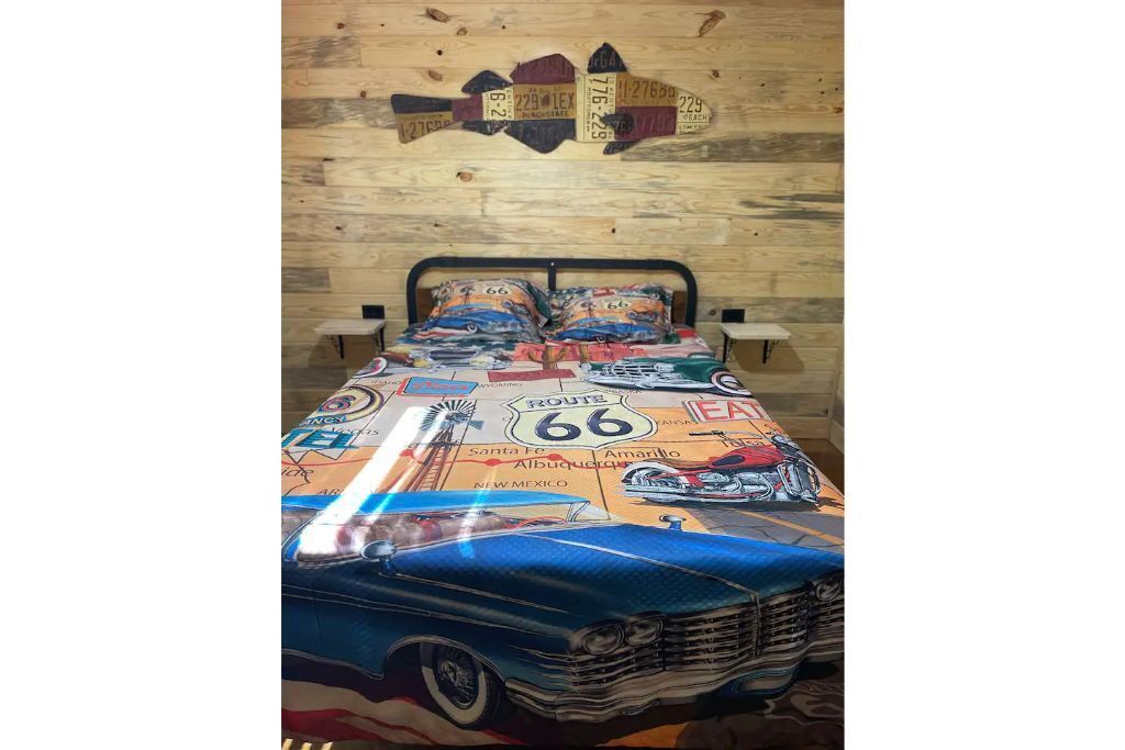 Un lit avec une couvre-lit sur le thème de la Route 66, un mur en bois agrémenté d’éléments décoratifs et une petite table de chevet.