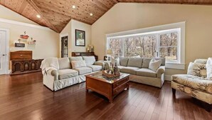 A spacious living room with wooden flooring, a high ceiling with wooden beams, and a large window offering a view of trees. - Perfect retreat for multi-generational families! Sat-Saturday lakeside rental. (Three Oaks)