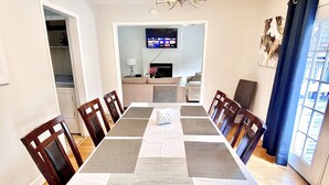 Una sala da pranzo con un lungo tavolo, sedie, una TV sullo sfondo e una finestra con tende.