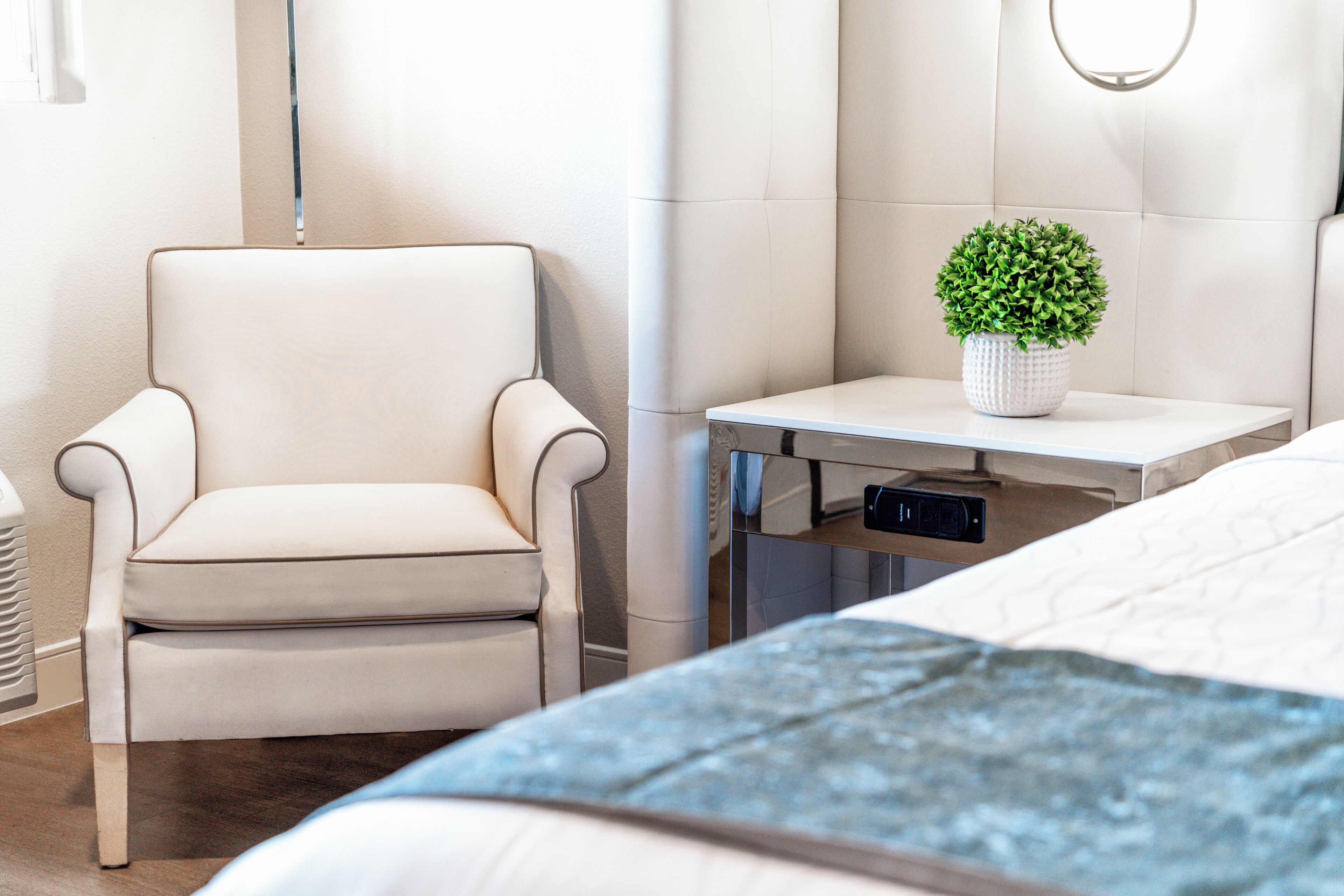 A modern bedroom with a bed, a chair, a bedside table with a plant, and a wall-mounted light fixture.