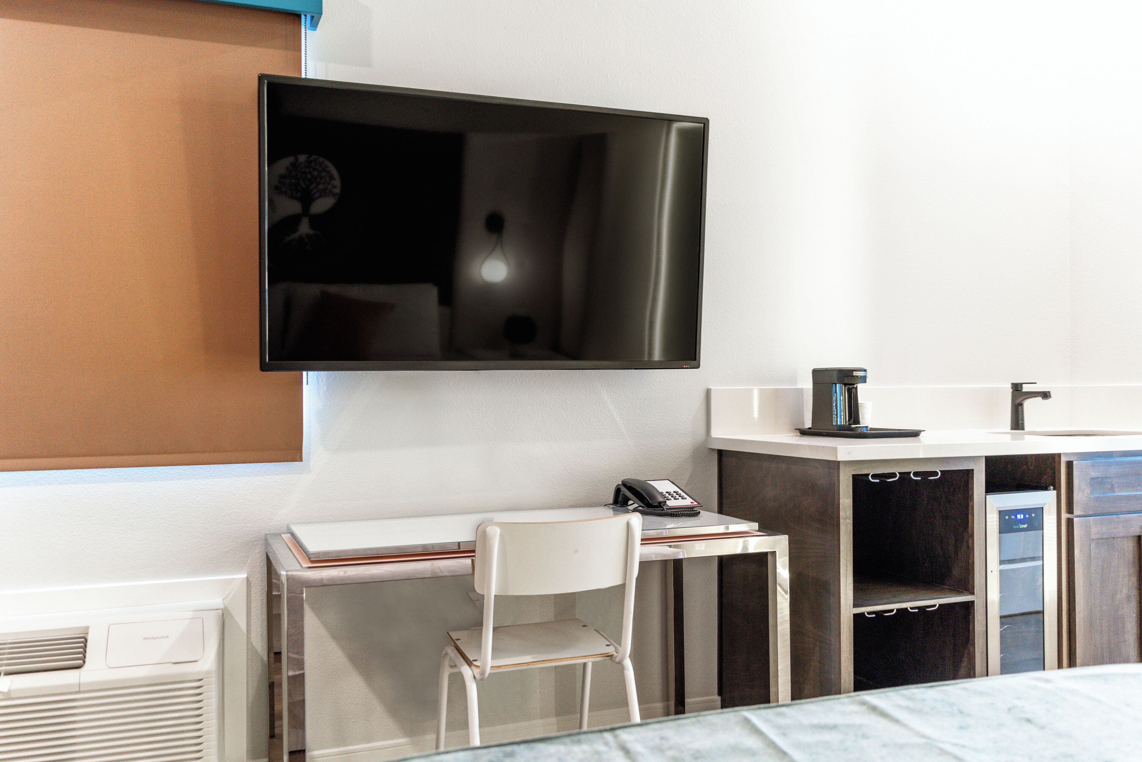 A hotel room with a flat-screen TV mounted on the wall, a desk with a chair, a coffee maker, and a built-in mini-fridge.