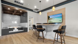 A modern kitchen with a wooden island, bar stools, and a large TV screen displaying a beach scene. - Aspire Inn Studios & Spas (Bellflower)