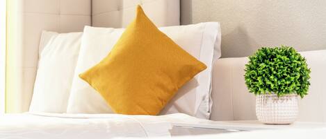 A neatly made bed with white bedding and a mustard-colored pillow, a small potted plant on a white bedside table, and a white headboard.