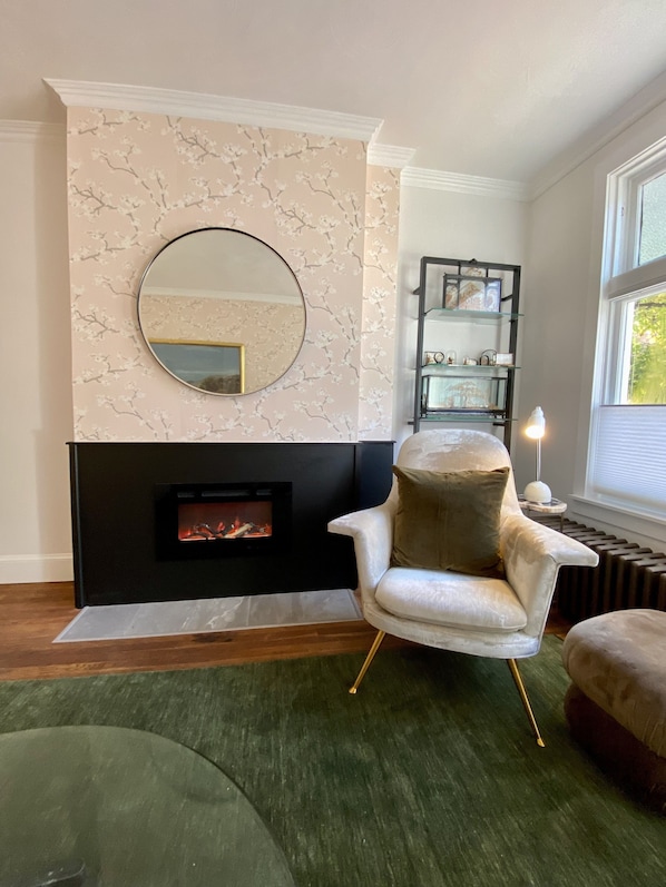 A living room with a fireplace, a round mirror, a white armchair, and a bookshelf.