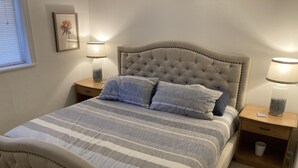 Un letto rifatto con una coperta a righe, due cuscini e una testiera imbottita. Sono presenti due comodini con lampade e un piccolo quadro appeso al muro.