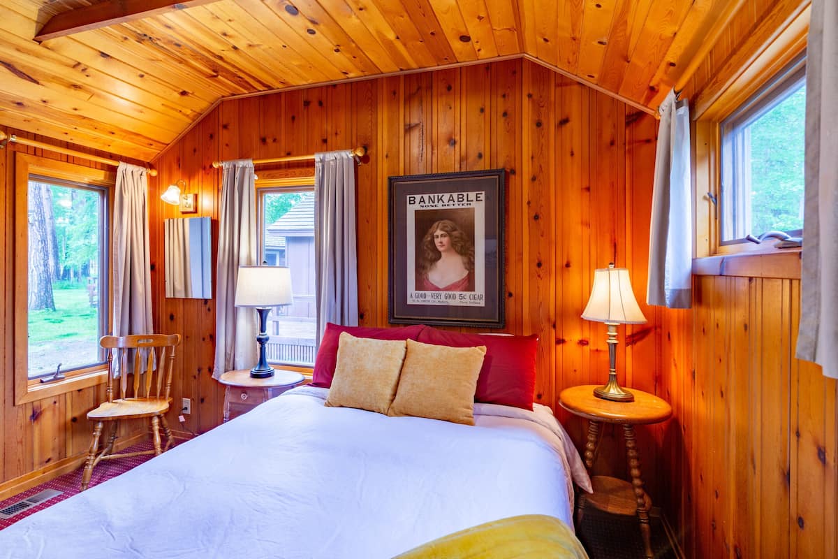 A wooden-paneled room with a bed, two bedside tables, a chair, and a poster on the wall.