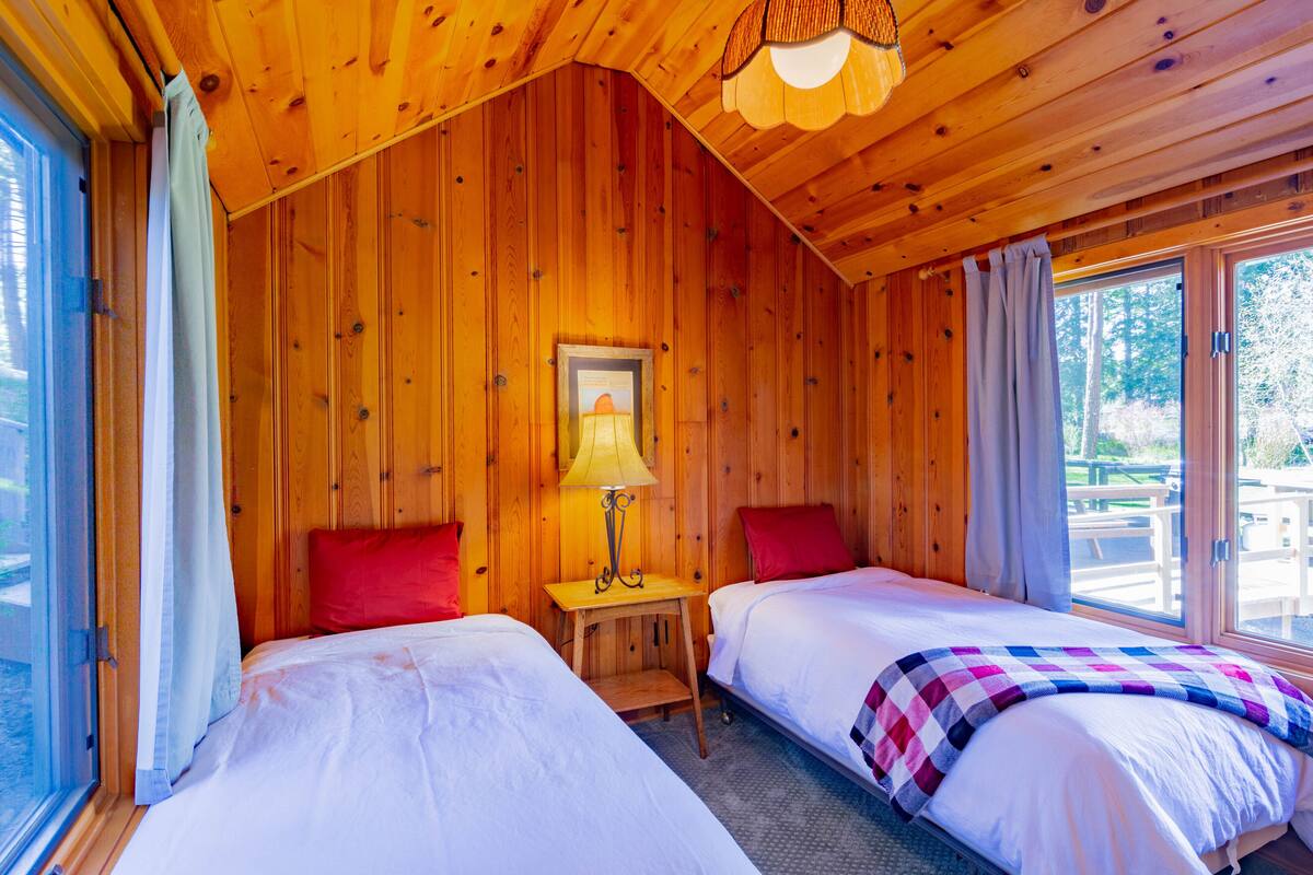 A wooden cabin room with two beds, a table with a lamp, and a window with curtains.