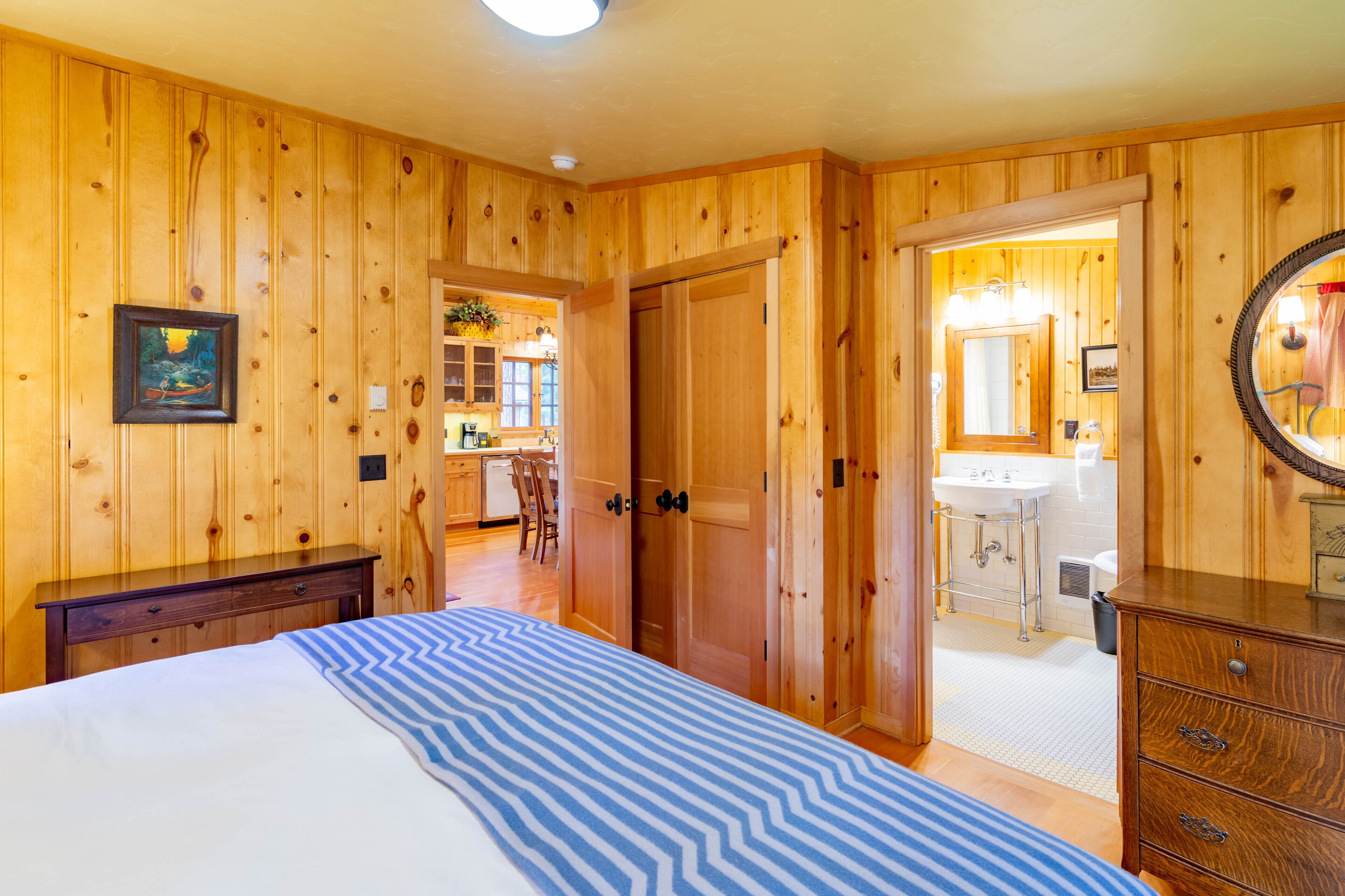 Una habitación con paneles de madera, una cama, un aparador y una puerta que da a un baño con lavabo y espejo.