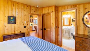 Una habitación con paneles de madera, una cama, un aparador y una puerta que da a un baño con lavabo y espejo.