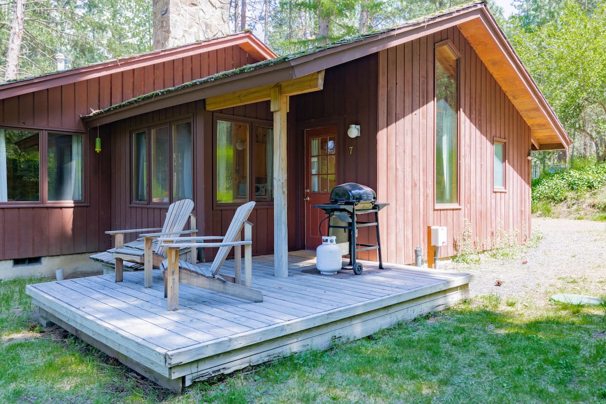 A wooden cabin with a porch, two chairs, a grill, and a gas tank.