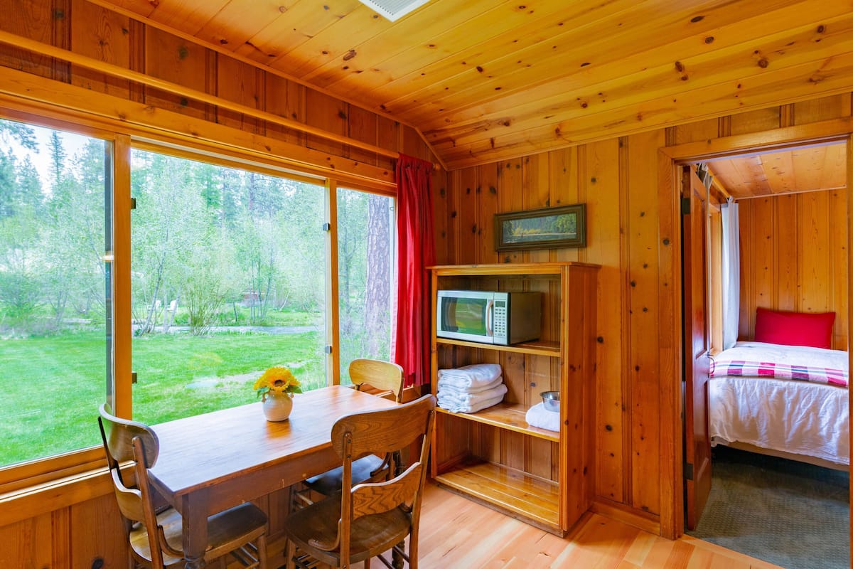 A wooden cabin interior with a dining table, chairs, a microwave, a bed, and a view of a forest.