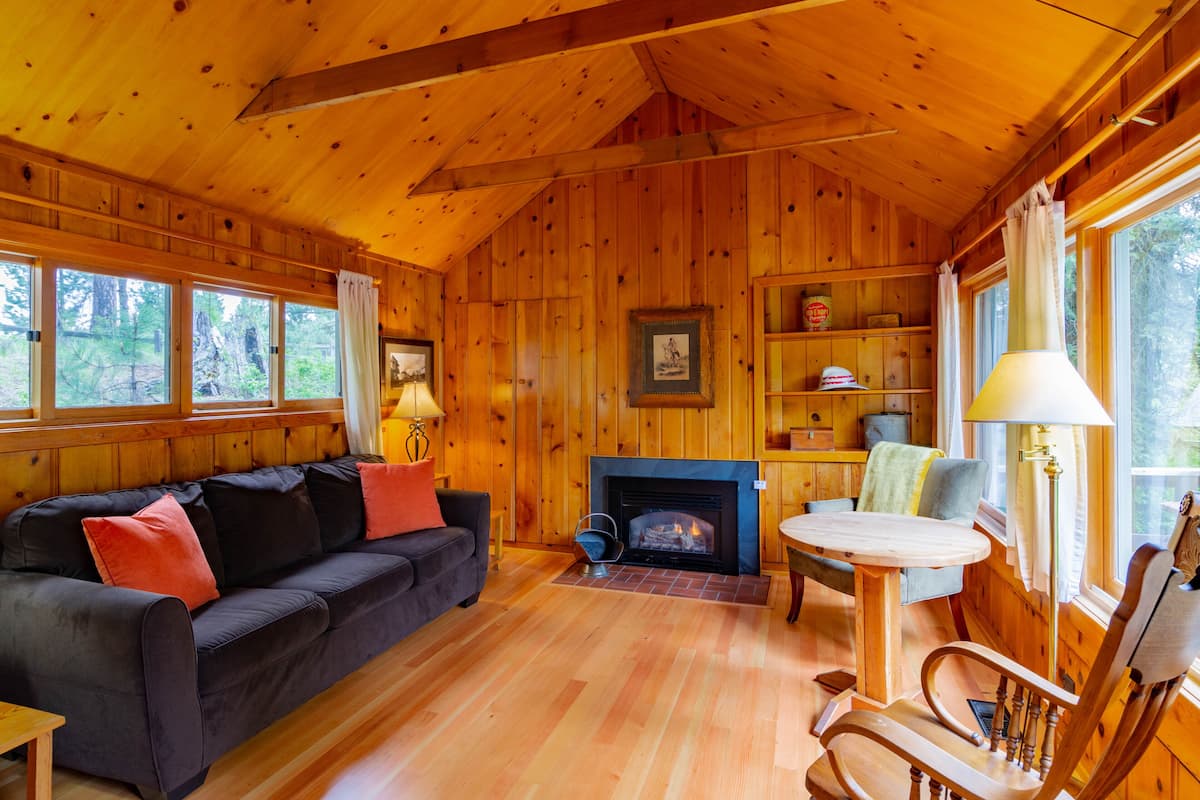 A cozy cabin interior with a wooden floor, a fireplace, a sofa, a dining table, and a chair.