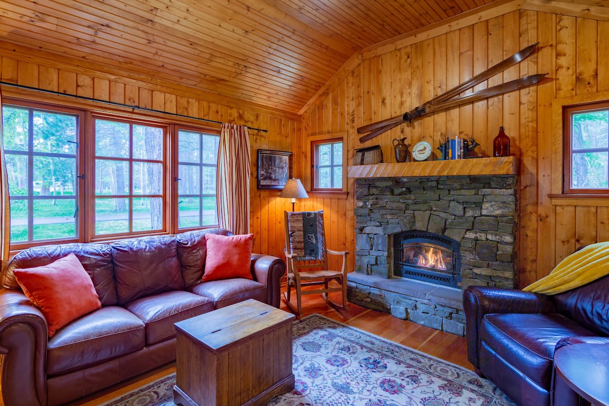 A wooden cabin interior with a stone fireplace, leather sofas, and large windows.