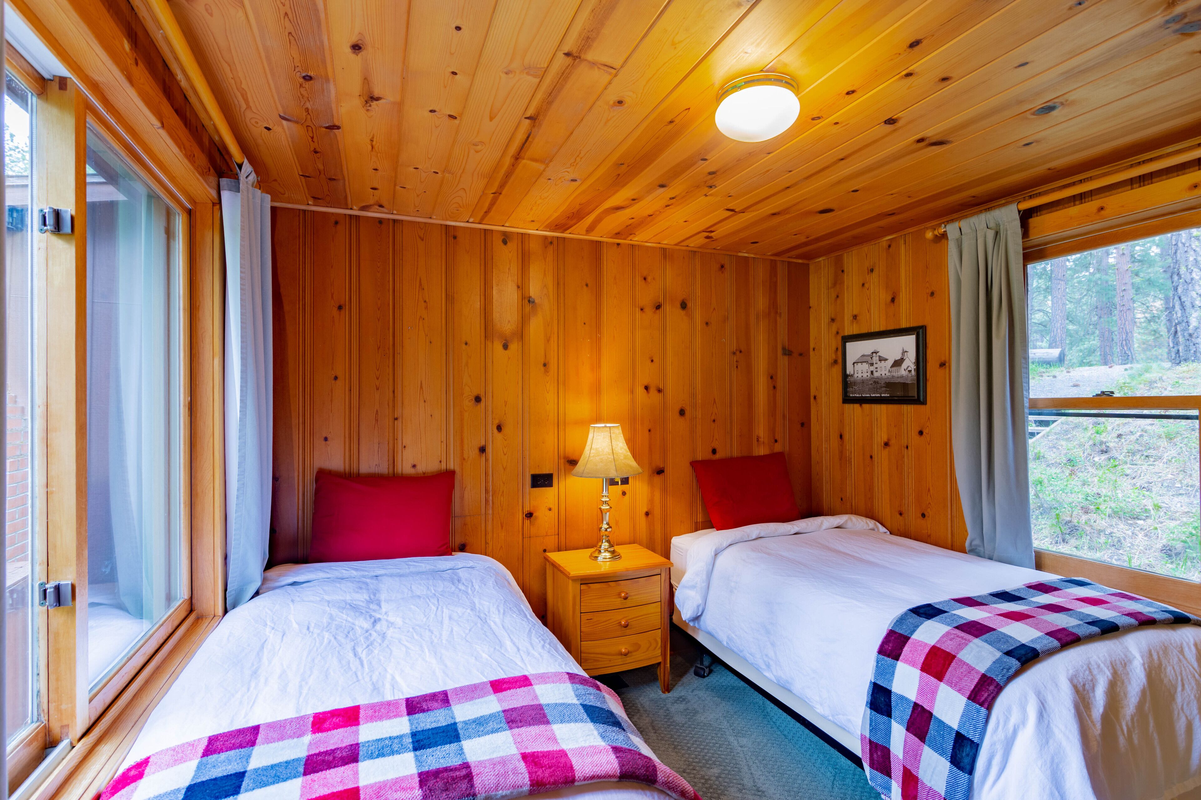 A wooden cabin room with two beds, a nightstand with a lamp, and a window with a view of trees.