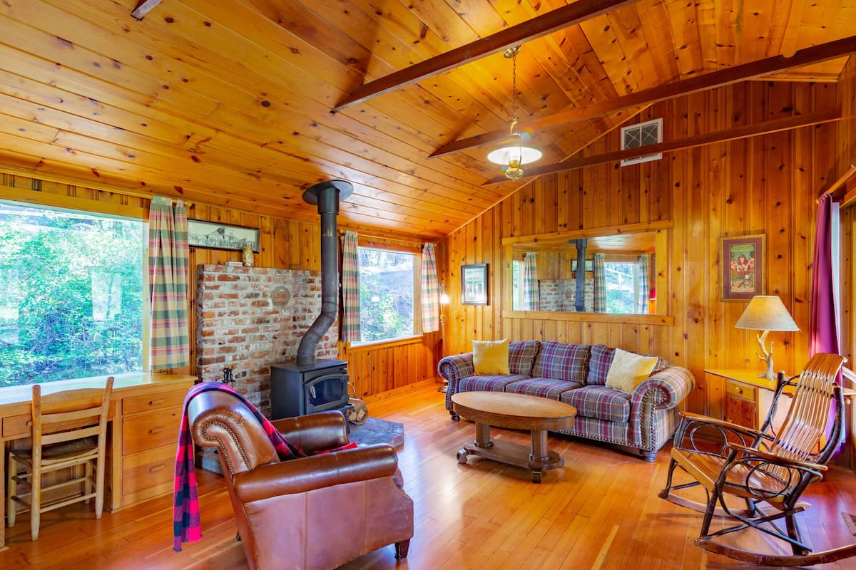 A cozy cabin interior with a wooden interior, a fireplace, a sofa, a rocking chair, and a dining area.