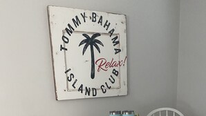 Une table ronde blanche, accompagnée de deux chaises blanches, un panneau indiquant « Tommy Bahama Island Club – Détendez-vous ! » et deux verres bleus posés sur la table.