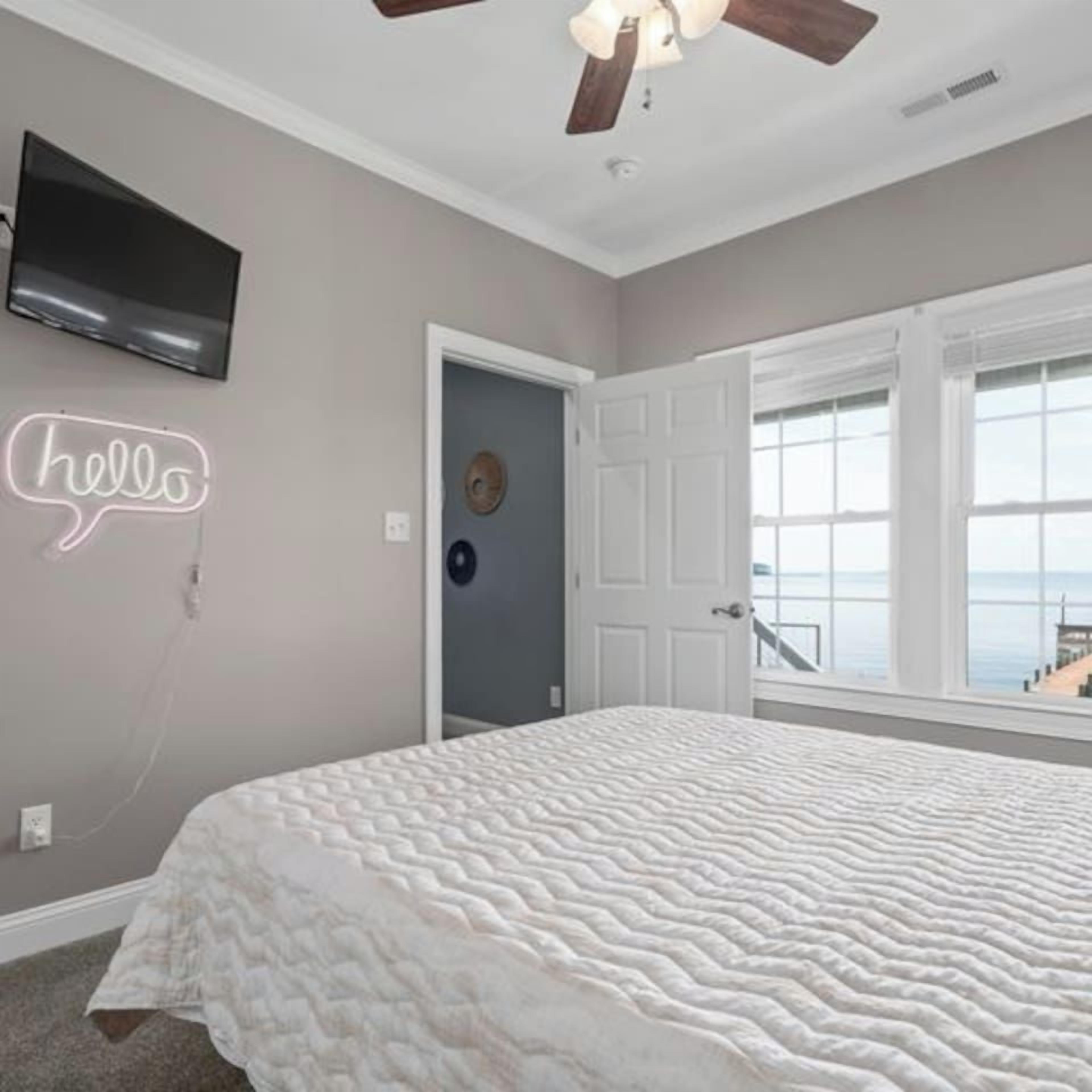 Un dormitorio con cama, ventilador de techo, televisión, un letrero de neón y vista al mar.
