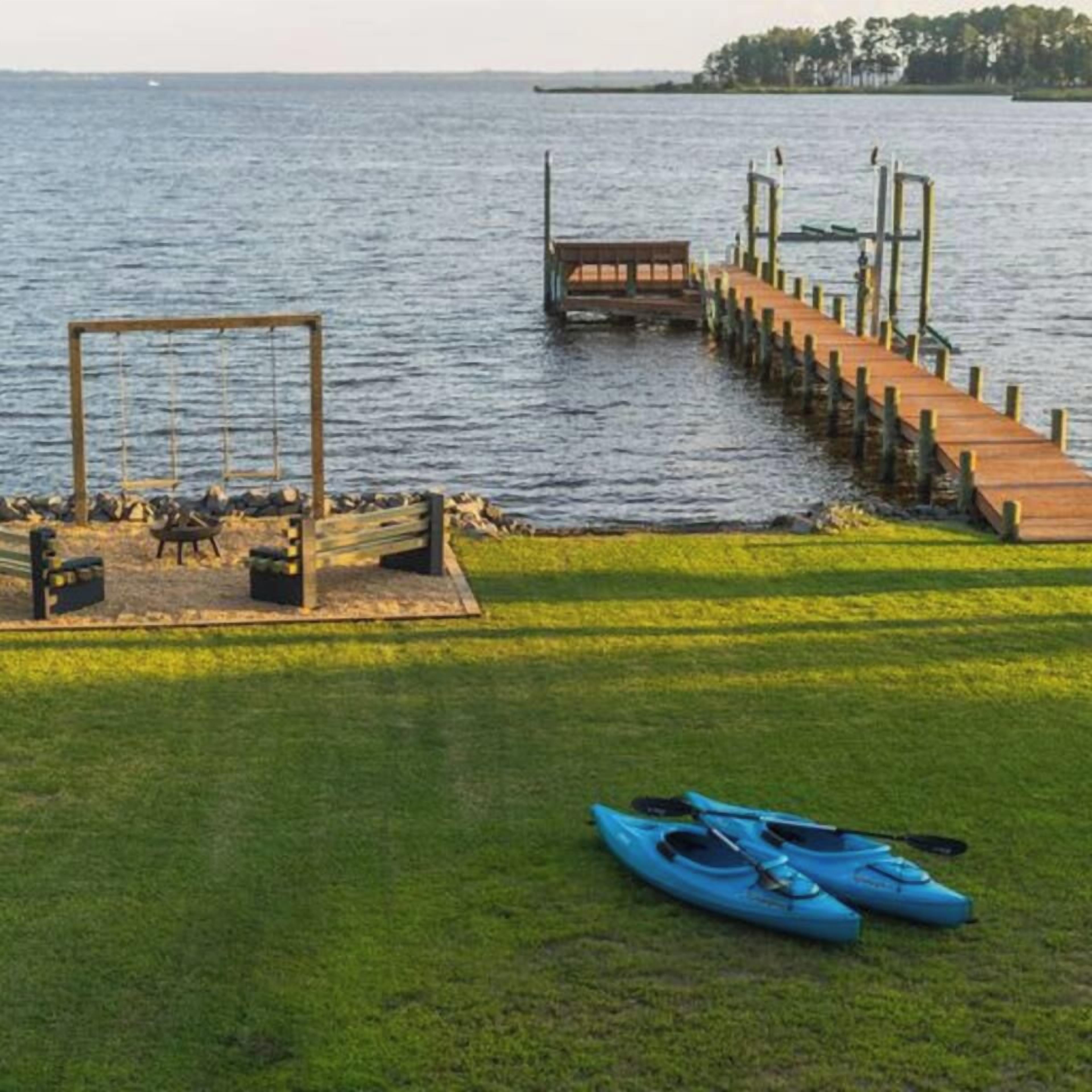 Une jetée en bois s’étendant sur une étendue d’eau calme, une zone herbeuse avec des kayaks, et une rive lointaine.
