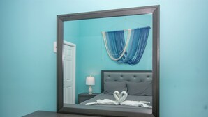 A bedroom with a large mirror, a bed with grey bedding, a bedside table with a lamp, and a blue curtain. - Brand new home in lively West End- Coral Cove (Roatan)
