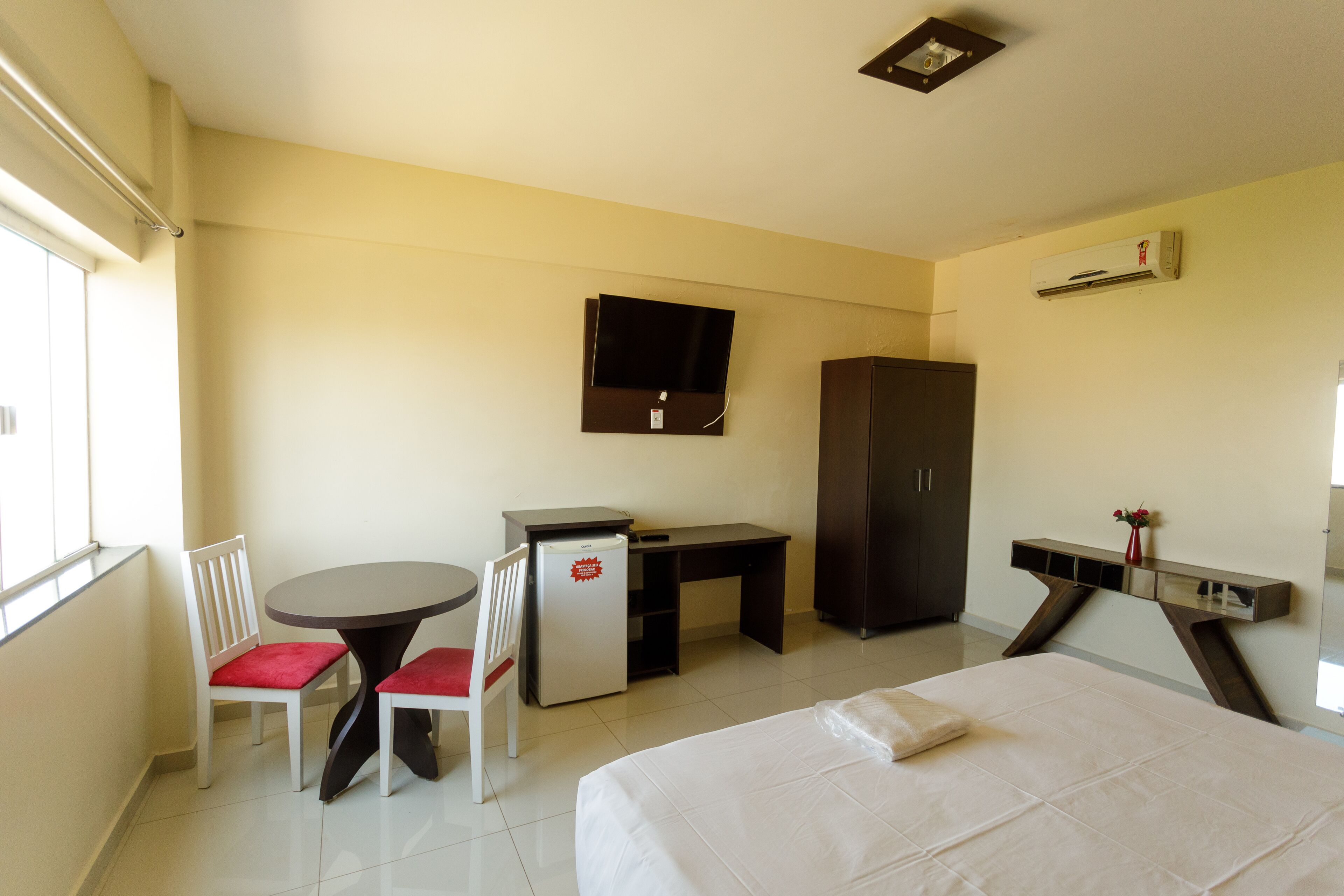 A hotel room with a bed, a desk, a chair, a small refrigerator, a television, and a wardrobe.