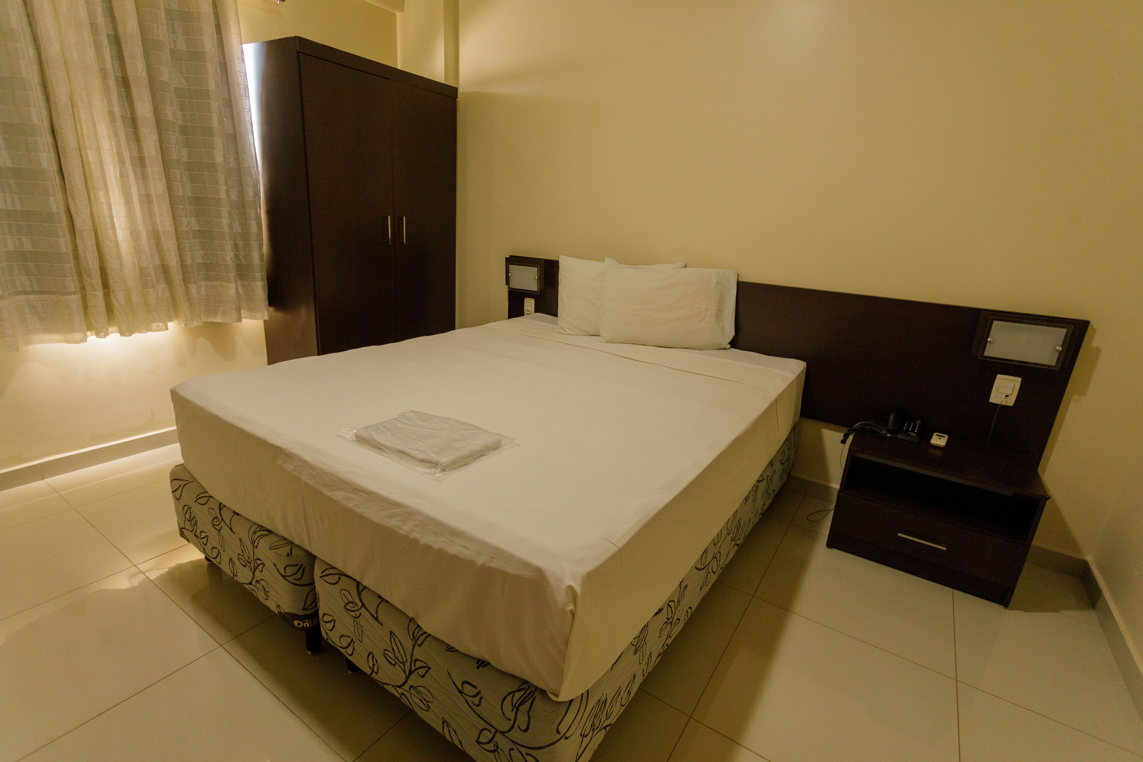 Cama bem arrumada com colcha branca e toalha dobrada, guarda-roupa de madeira escura, mesa de cabeceira com abajur e uma pequena cômoda.