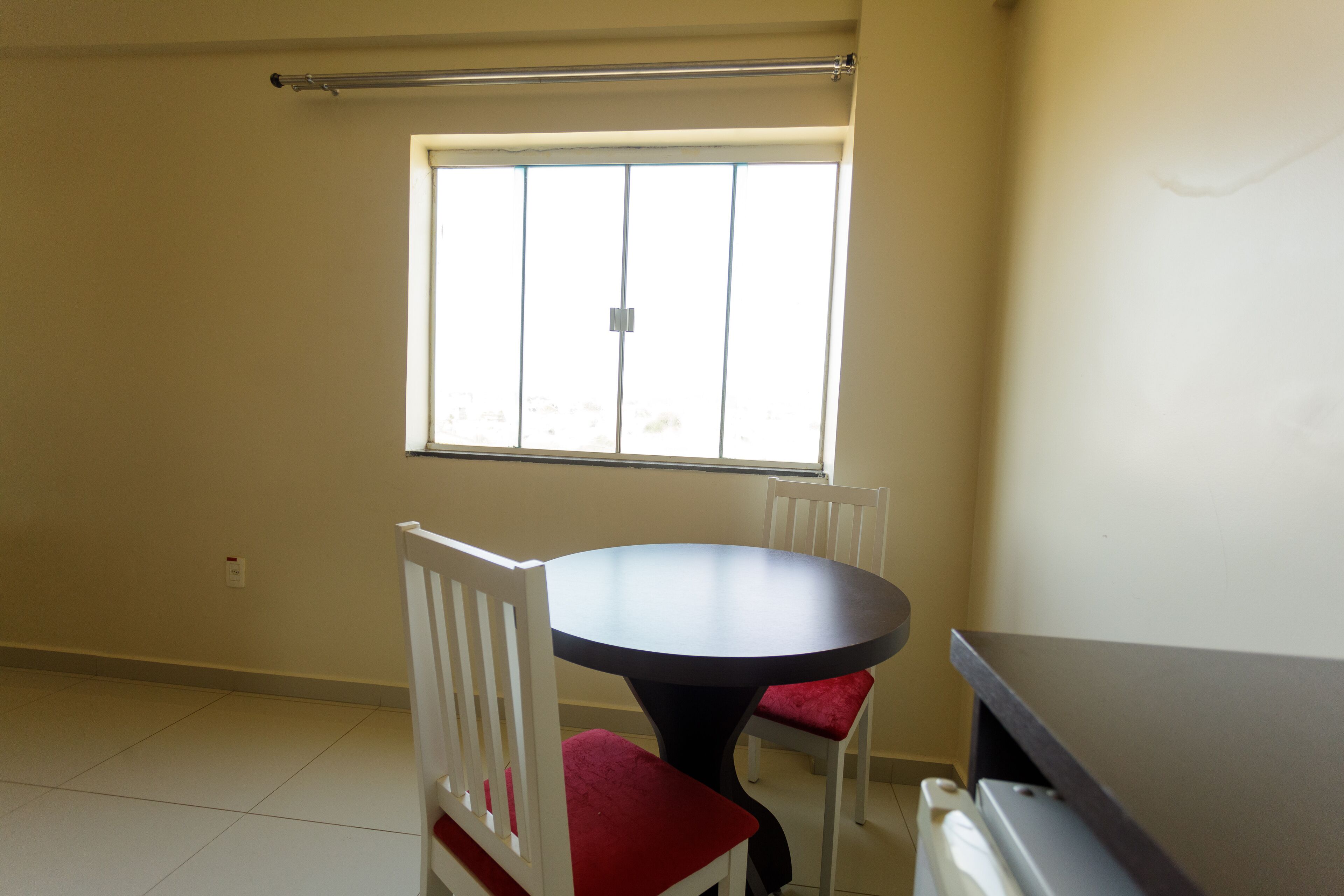 Une pièce avec une table ronde, deux chaises blanches avec des coussins rouges, une fenêtre avec des stores et un radiateur.