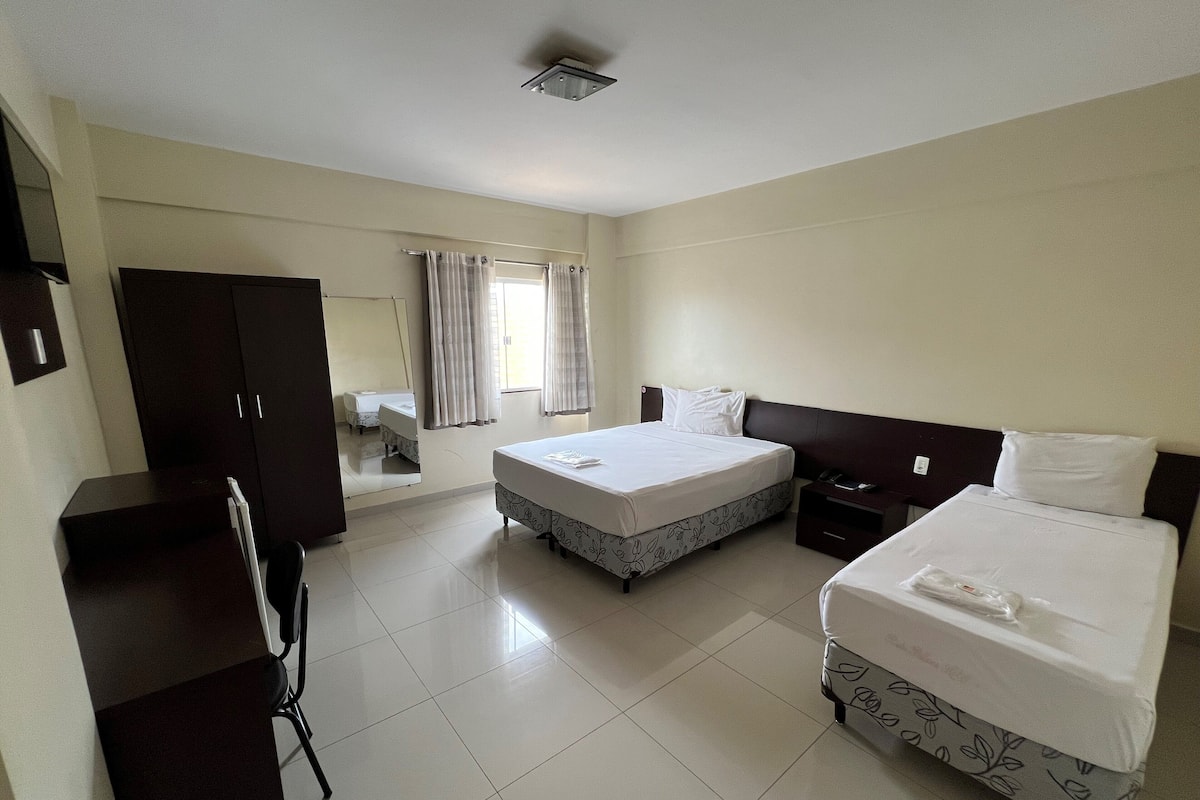 Quarto de hotel com duas camas, uma escrivaninha, um guarda-roupa e uma casa de banho visível através de uma porta.