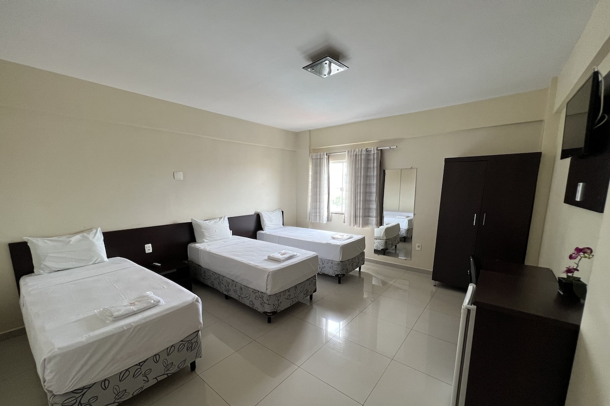 Quarto de hotel com três camas, um guarda-roupa escuro, uma pequena escrivaninha e um espelho.