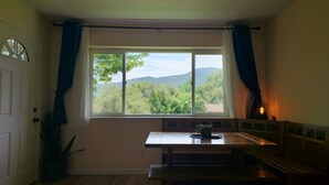 Una habitación con un banco de madera, una mesa, una ventana con cortinas y vistas a árboles y colinas.
