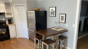 A kitchen with a stainless steel refrigerator, a wooden table with two stools, and a basket on top of the fridge. There are framed pictures on the wall and a basket on the floor. - Visit Monticello, Wineries, UVA & More (Troy)