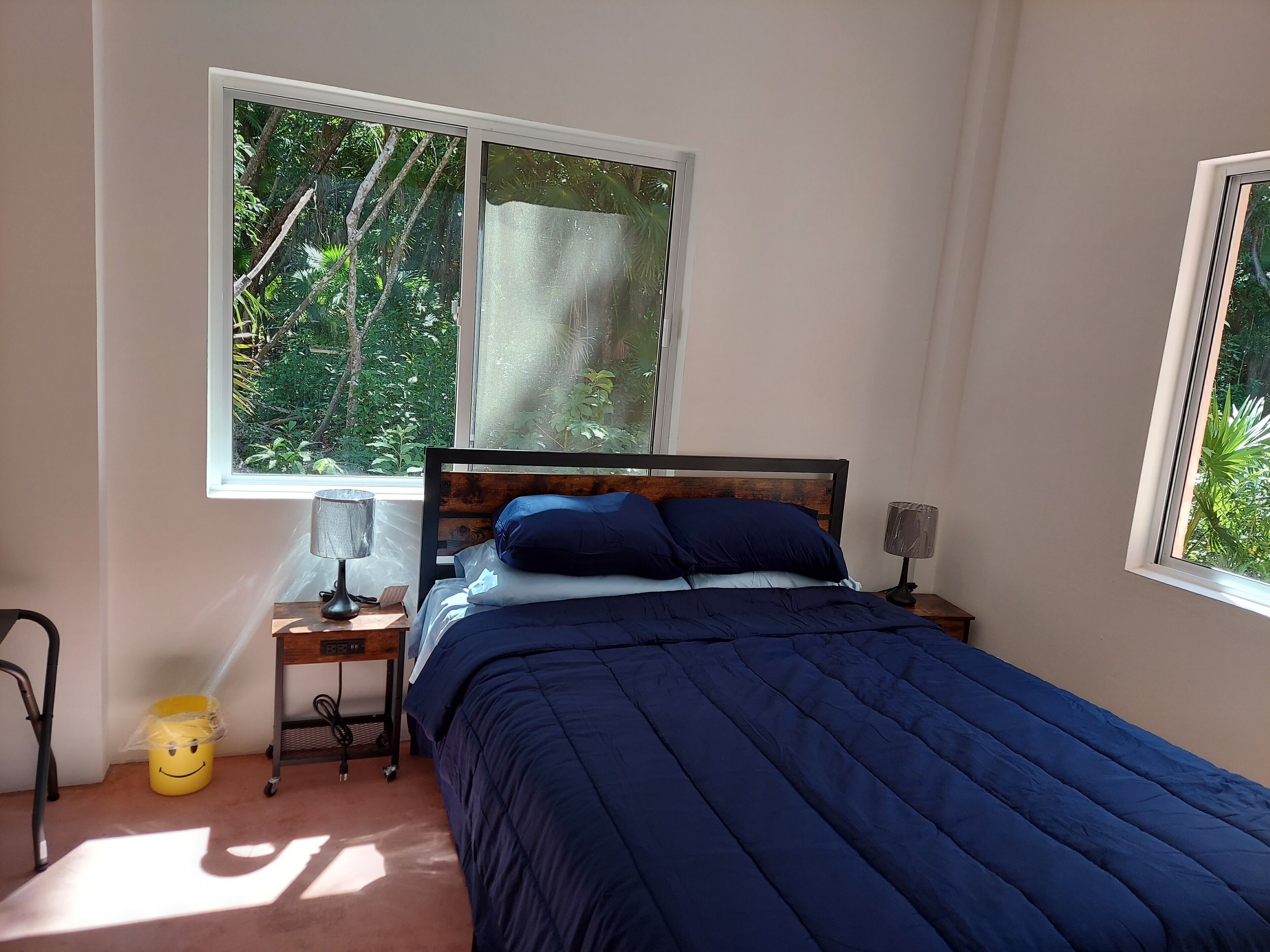 A bedroom with a bed, two bedside tables, a window, and a small trash bin.