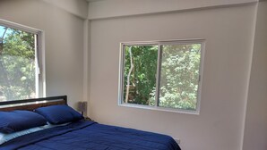 Un dormitorio con una cama, dos ventanas, una mesita de noche, una lámpara y un cubo de basura con una carita sonriente.