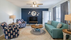 Ein Wohnzimmer mit Deckenventilator, Fernseher, blauem Sofa, gemustertem Sessel und Couchtisch.