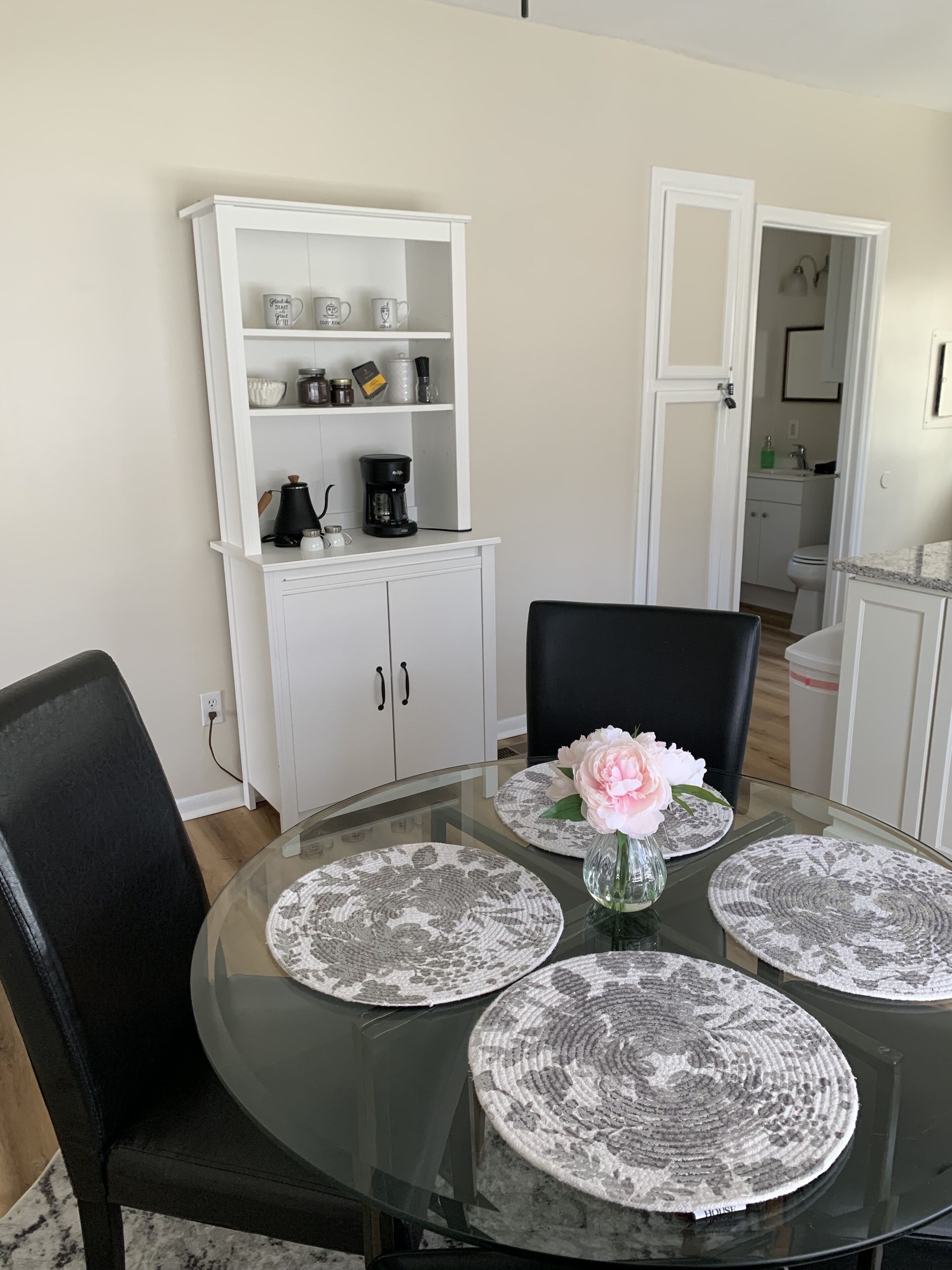 A dining area with a glass table, black chairs, and a white cabinet with kitchen items.