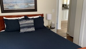 A bedroom with a wooden bed, a framed landscape painting, a bedside lamp, and a view of the kitchen through an open door. - Jo Mac Cottage- Quiet Home near UNC Chapel Hill (Chapel Hill)