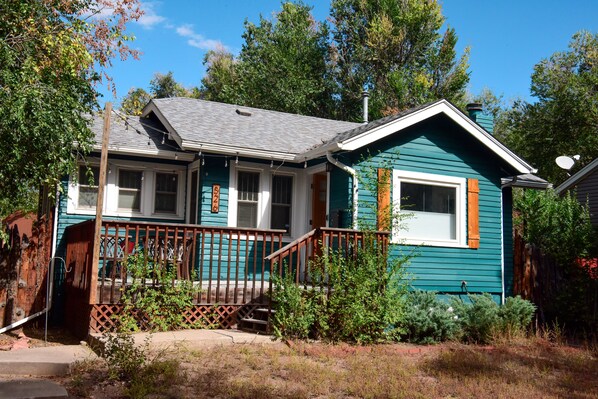 Charming single-story house with a steeply pitched roof and front porch, surrounded by a wooden fence and a garden with various shrubs and plants.