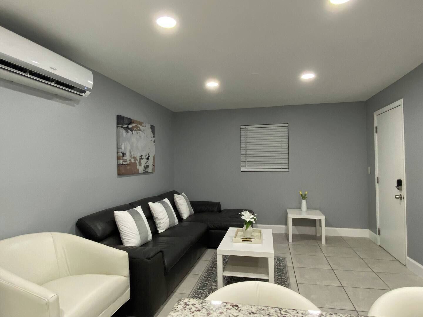 A modern living room with a black sofa, white armchairs, a small white table, and a grey wall with a window and an air conditioning unit.