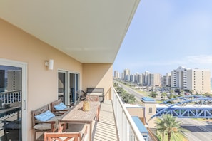 Uma varanda com mesa e cadeiras de madeira, um sofá com almofadas e vista para um horizonte urbano com edifícios altos e uma ponte.