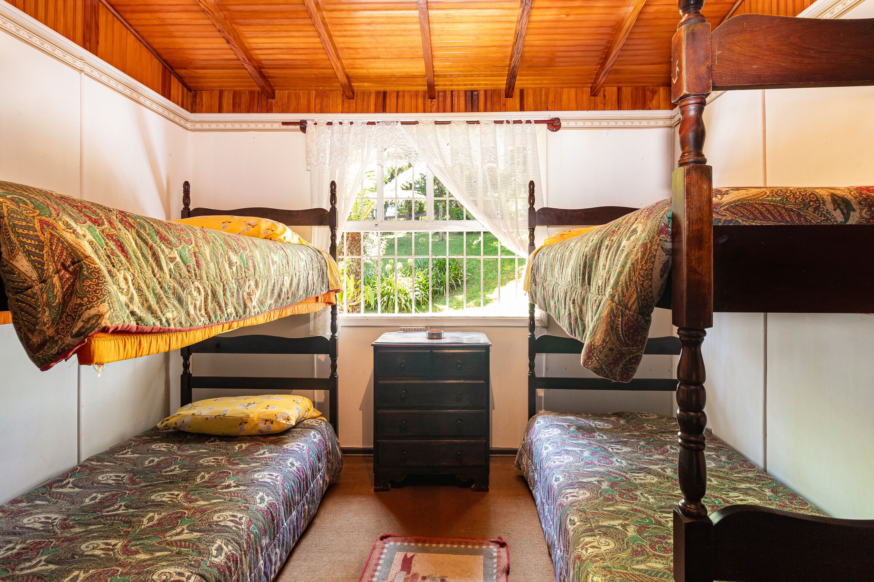 Quarto pequeno e acolhedor com duas beliches, uma cômoda de madeira e uma janela com vista para o verde.