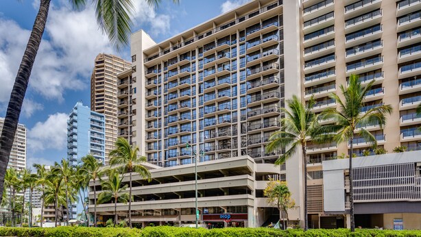 A multi-story hotel with a modern design, featuring a central atrium with a shopping area, surrounded by palm trees and other buildings.
