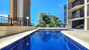A swimming pool with a clear blue water, surrounded by a low stone wall, and overlooking tall buildings and palm trees.