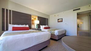 A hotel room with two beds, a nightstand with a lamp, a wall-mounted TV, and a door leading to another room.
