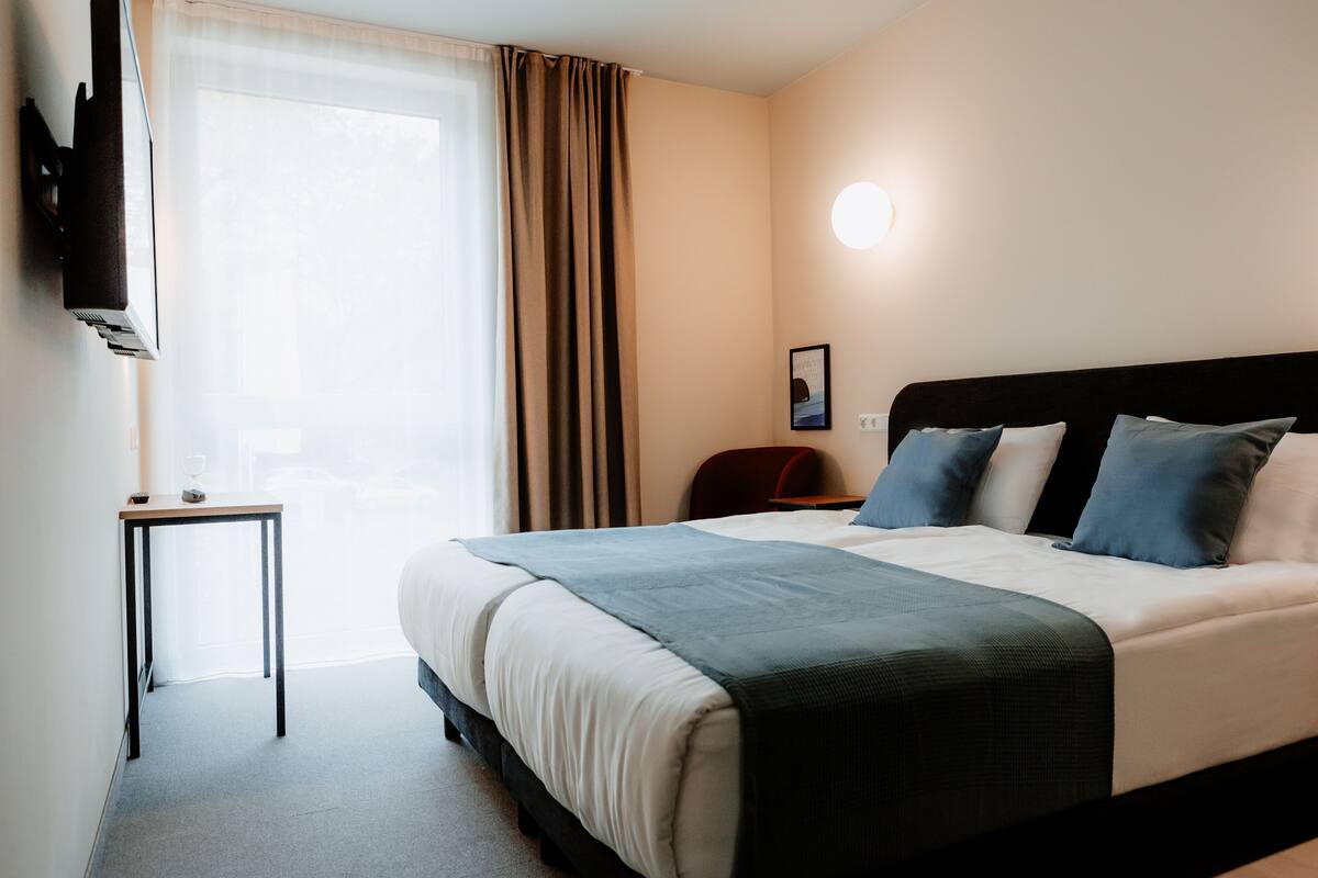Une chambre d’hôtel avec un lit, une table de chevet, un téléviseur fixé au mur et une fenêtre avec des rideaux.