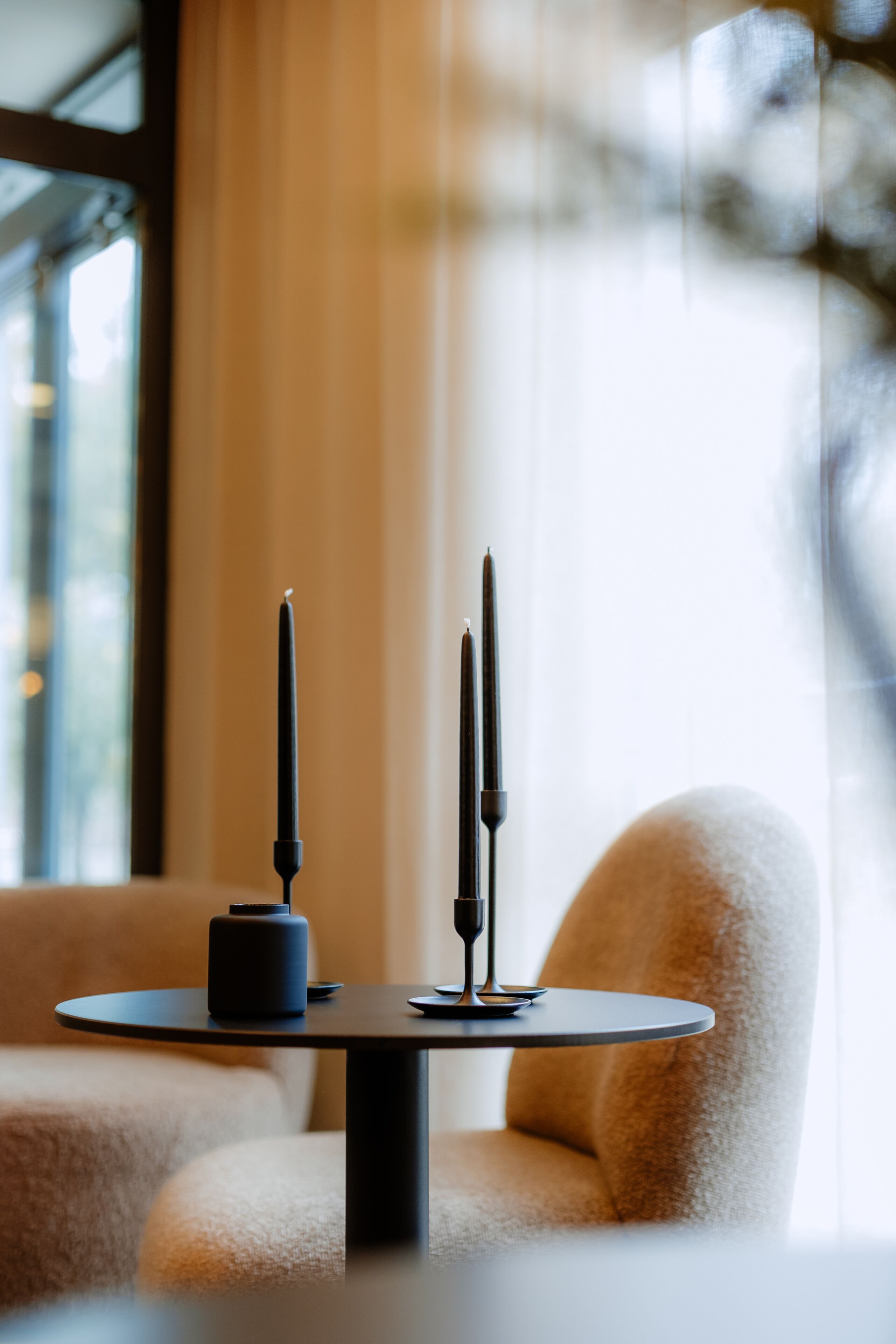 A round table with two black candles and a small black object, likely a candle holder, in a room with a large window and a soft beige chair.