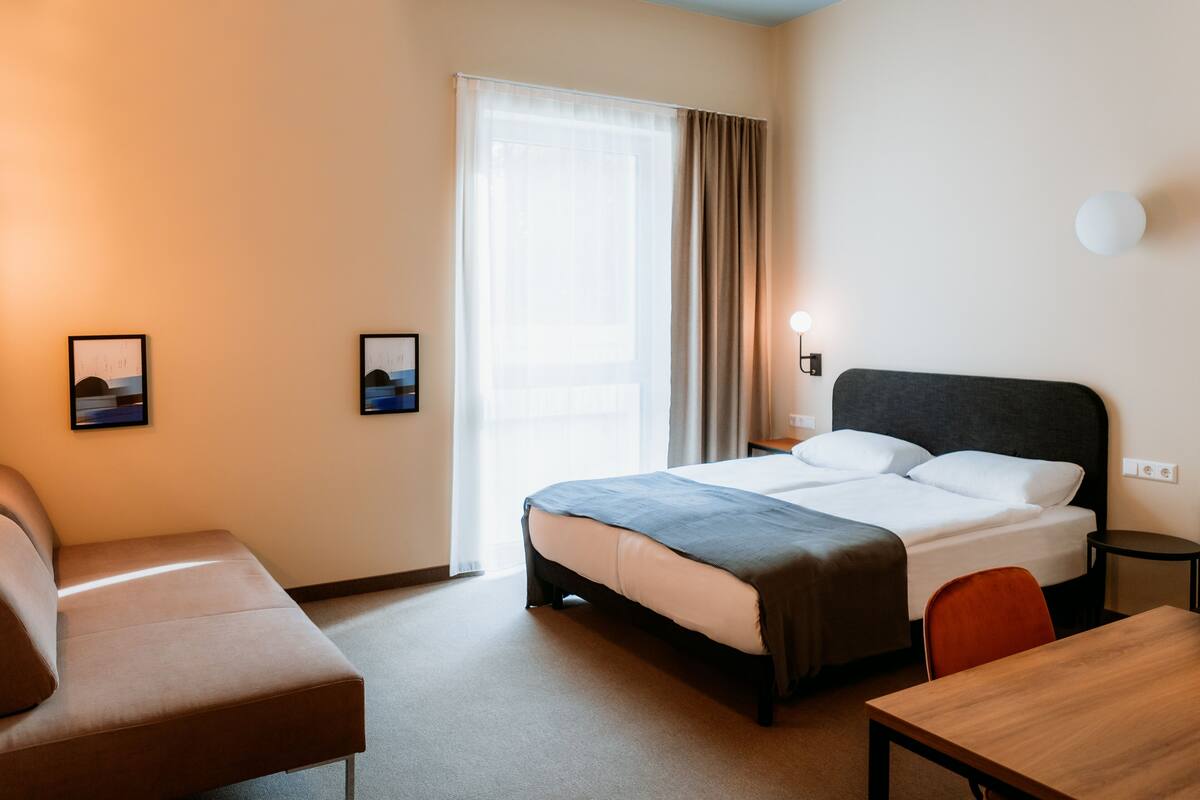 Une chambre d’hôtel comprenant un lit, un bureau, une chaise, un canapé, une fenêtre avec des rideaux et deux cadres photos fixés au mur.