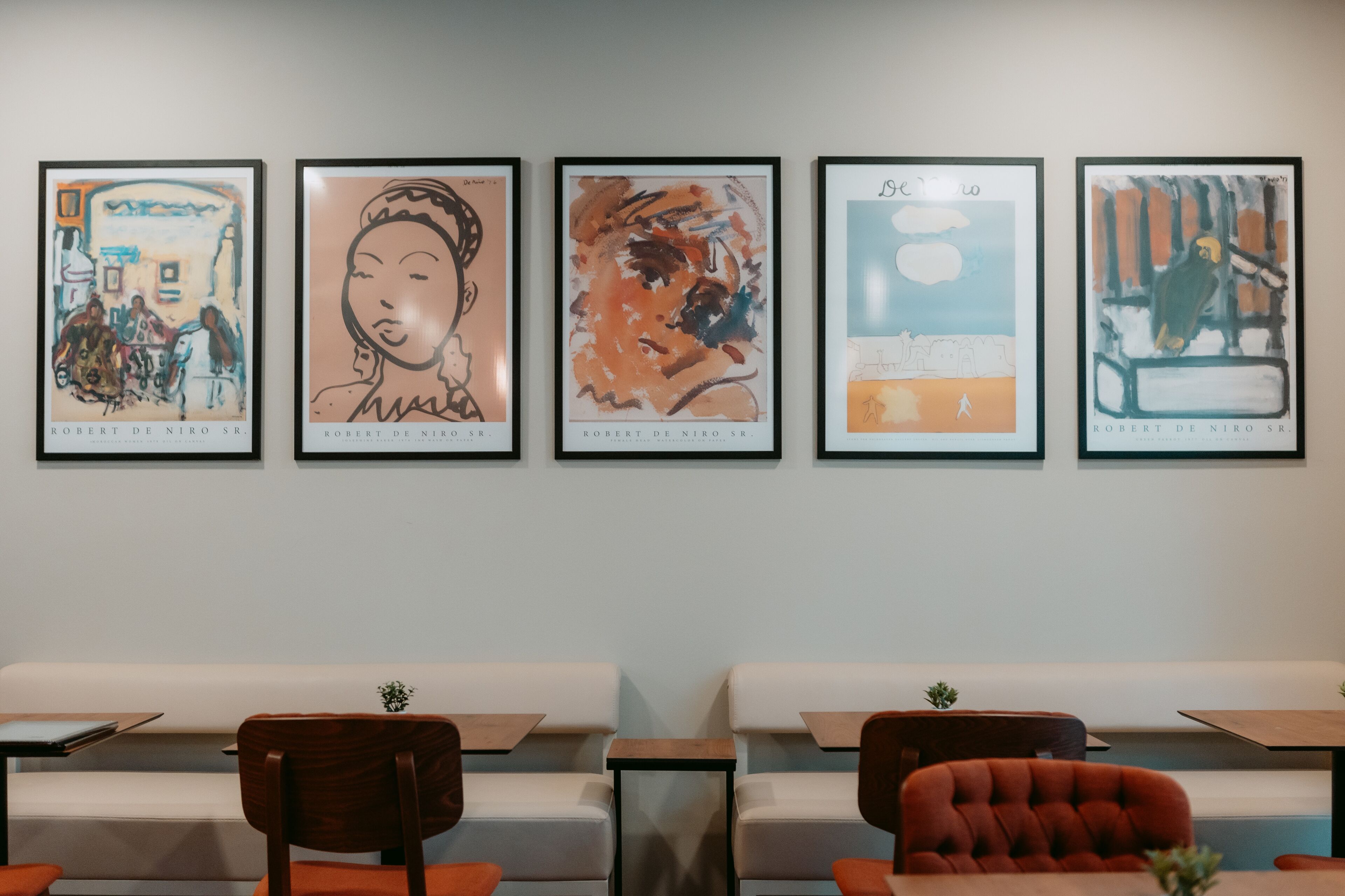 Eine Wand mit fünf gerahmten Kunstwerken von Robert De Niro Sr. und eine Sitzgelegenheit mit weißen Bänken und Holztischen.