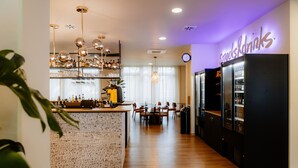 A modern kitchen with a bar area, including a refrigerator labeled "Snacks & Drinks," a dining table with chairs, and a chandelier. - Moods Hotel Dortmund (Dortmund)